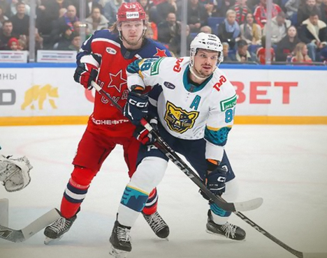 На изображении два хоккеиста на льду: один в красной форме с логотипом звезды, другой в белой форме с изображением снежного барса. Они находятся в движении, пытаясь завладеть шайбой. На заднем плане видно зрителей