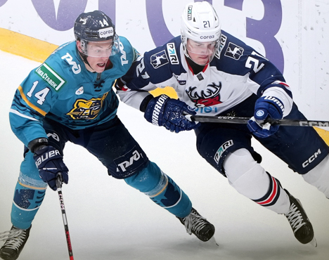 На изображении два хоккеиста в динамике на льду в момент игры. Один одет в форму бирюзового цвета, другой в тёмно-синюю с белыми элементами. Они сосредоточены на сопротивлении и держат клюшки в руках