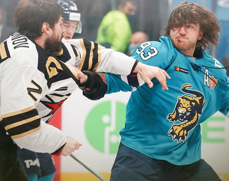 На изображении два хоккеиста, один в чёрной форме, другой в голубой, участвуют в потасовке на льду во время матча. На заднем плане видны зрители и еще один игрок в синей форме