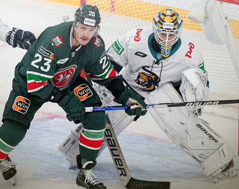 Хоккеист в зеленой форме с номером 23 держит клюшку, находясь перед вратарем в бело-синей форме и маске. Оба находятся на ледовой площадке во время игры